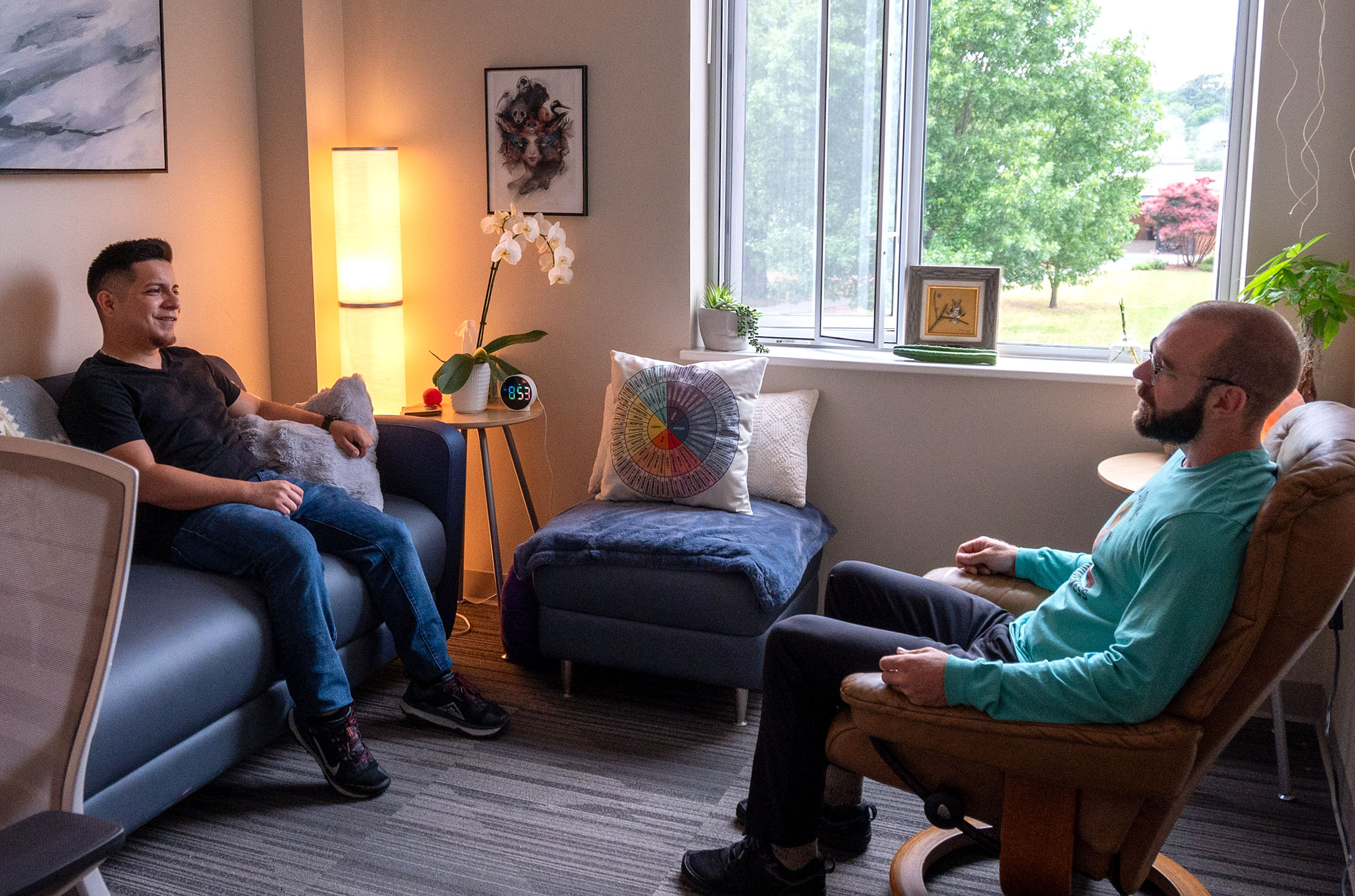 Two people sit facing each other in comfortable chairs, surrounded by soft lighting, plants, artwork and a large window overlooking trees, creating a welcoming and relaxed environment for conversation and support.