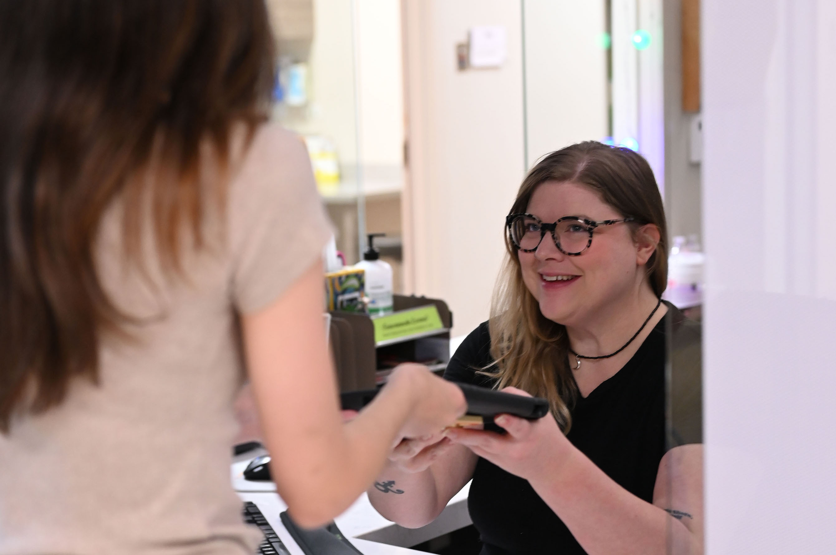 A young brunette woman working at the GYN (Women's) Clinic intake passes information to a female student to fill out before their appointment.